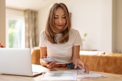 A woman is sitting at a wooden table, holding a smartphone and sorting through papers in a well-lit home office filled with natural light.