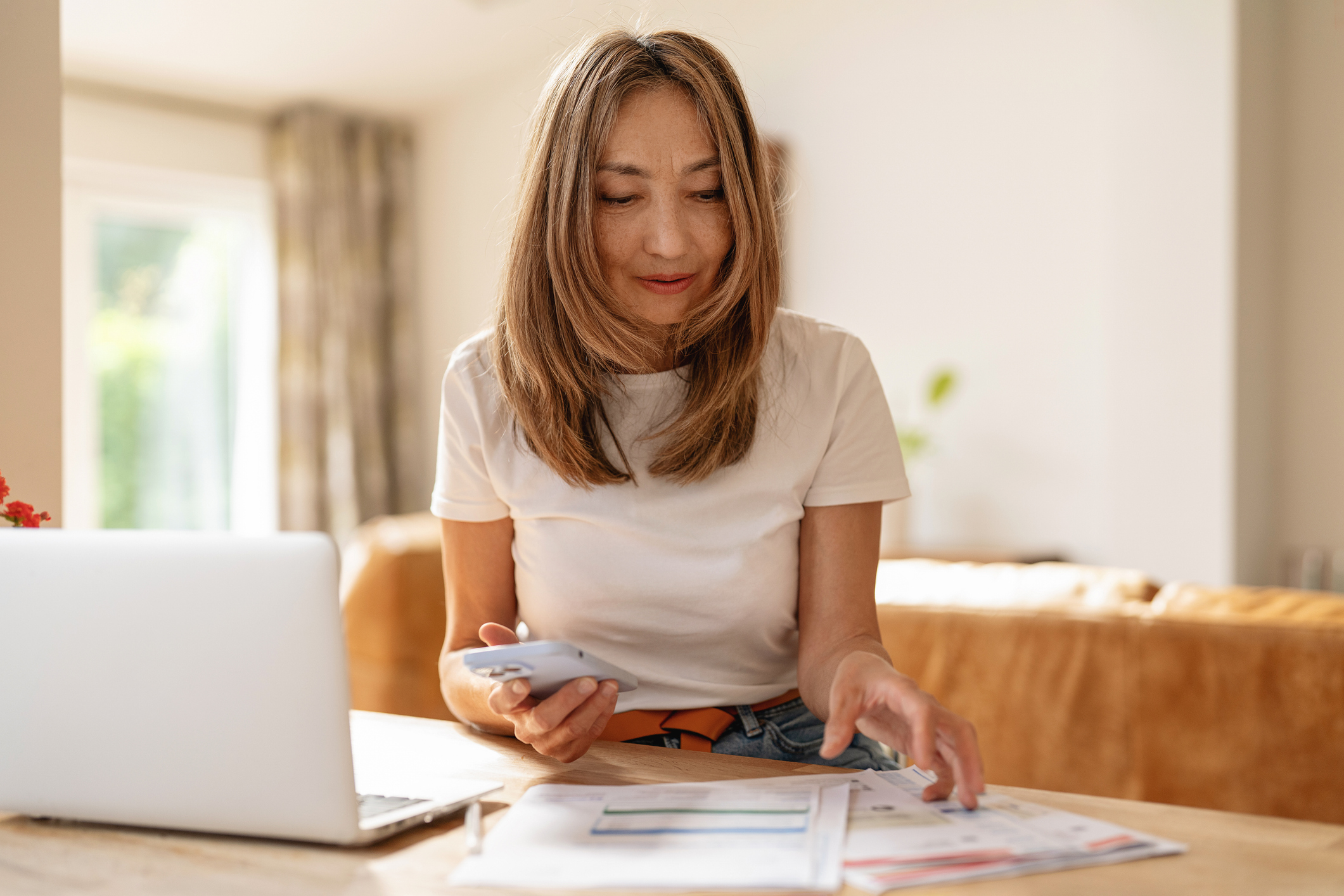 A woman is sitting at a wooden table, holding a smartphone and sorting through papers in a well-lit home office filled with natural light.